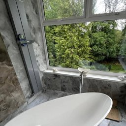 Łukasz - Nowoczesna, wolnostojąca wanna owalna z chromowaną baterią, ustawiona pod oknem z widokiem na zieleń, ściany i podłoga wyłożone płytkami imitującymi marmur w odcieniach szarości, obok wanny leżą...