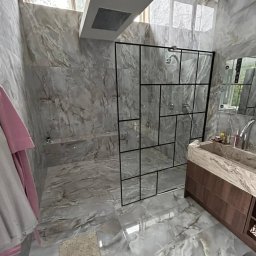 Łukasz - Nowoczesna łazienka wykończona płytkami imitującymi marmur, kabina prysznicowa z czarnymi profilami i geometrycznym podziałem szklanych tafli, drewniana szafka pod umywalką z marmurowym blatem.