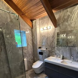 Łukasz - Nowoczesna łazienka z marmurowymi płytkami na ścianach i podłodze, drewnianym sufitem i podwójną umywalką z ciemnoszarą szafką, z widoczną kabiną prysznicową ze szklanymi drzwiami oraz toaletą.