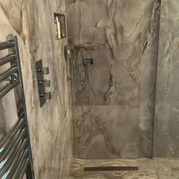 Łukasz - Nowoczesna kabina prysznicowa z odpływem liniowym, wykończona płytkami imitującymi marmur, z chromowaną armaturą i grzejnikiem drabinkowym.