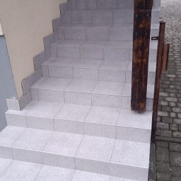 Łukasz - Zewnętrzne schody wyłożone jasnoszarymi płytkami, z drewnianą barierką po prawej stronie, widoczny pojemnik z wodą i narzędzia na dole.