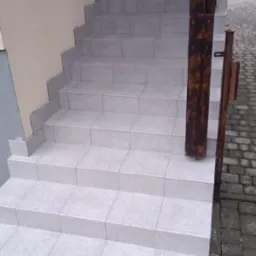 Zewnętrzne schody wyłożone jasnoszarymi płytkami, z drewnianą barierką po prawej stronie, widoczny pojemnik z wodą i narzędzia na dole.