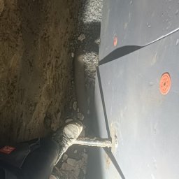 Łukasz - Instalacja rury w wykopie przy ścianie w Łańcucie, widoczne elementy mocujące i kamieniste podłoże.