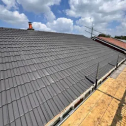 Nowy, szary dach z dachówki ceramicznej z widocznymi rusztowaniami i fragmentem nieba z chmurami.