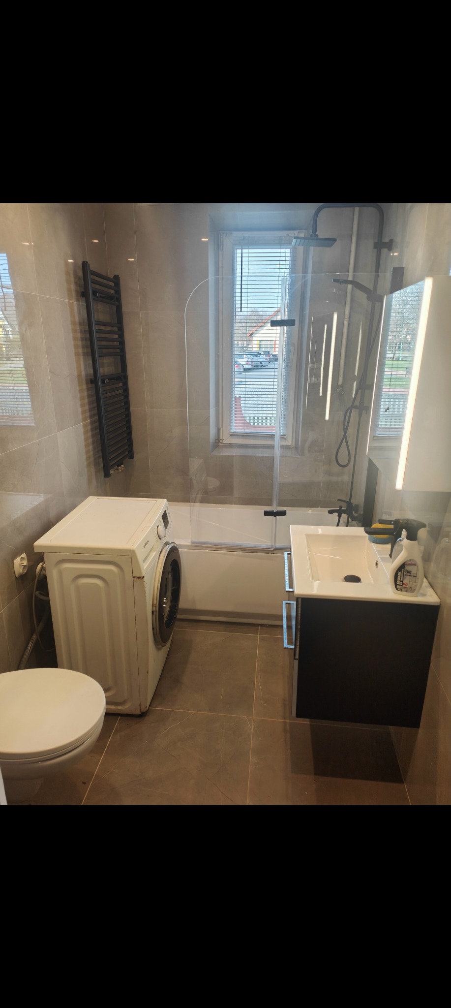 Ustawna łazienka z pralką obok toalety, kabiną prysznicową nad wanną, umywalką z szafką w kolorze czarnym, grzejnikiem drabinkowym i oknem z żaluzjami.