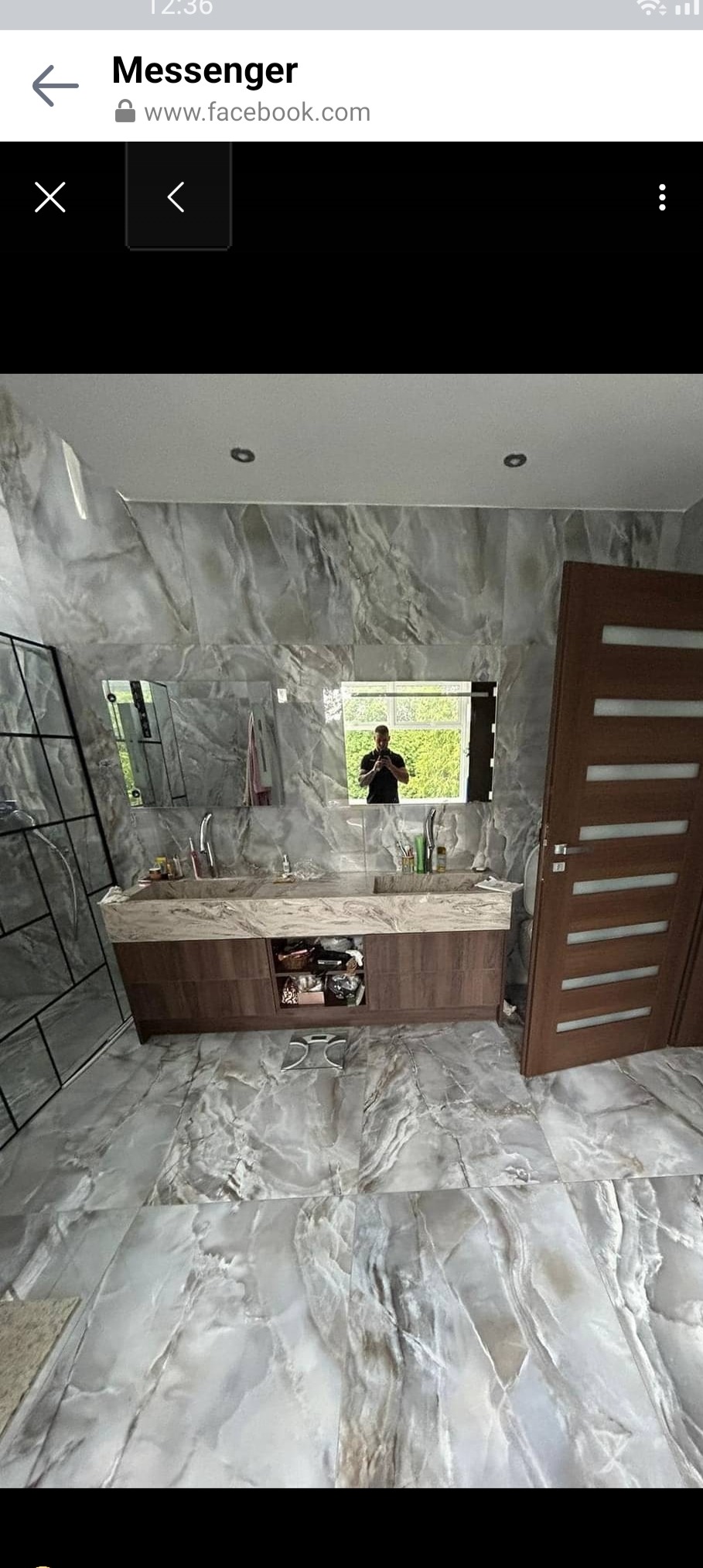 Wykończona łazienka z marmurowymi płytkami na ścianach i podłodze, drewnianą szafką pod umywalką z marmurowym blatem i dużym lustrem, w którym odbija się osoba robiąca zdjęcie.