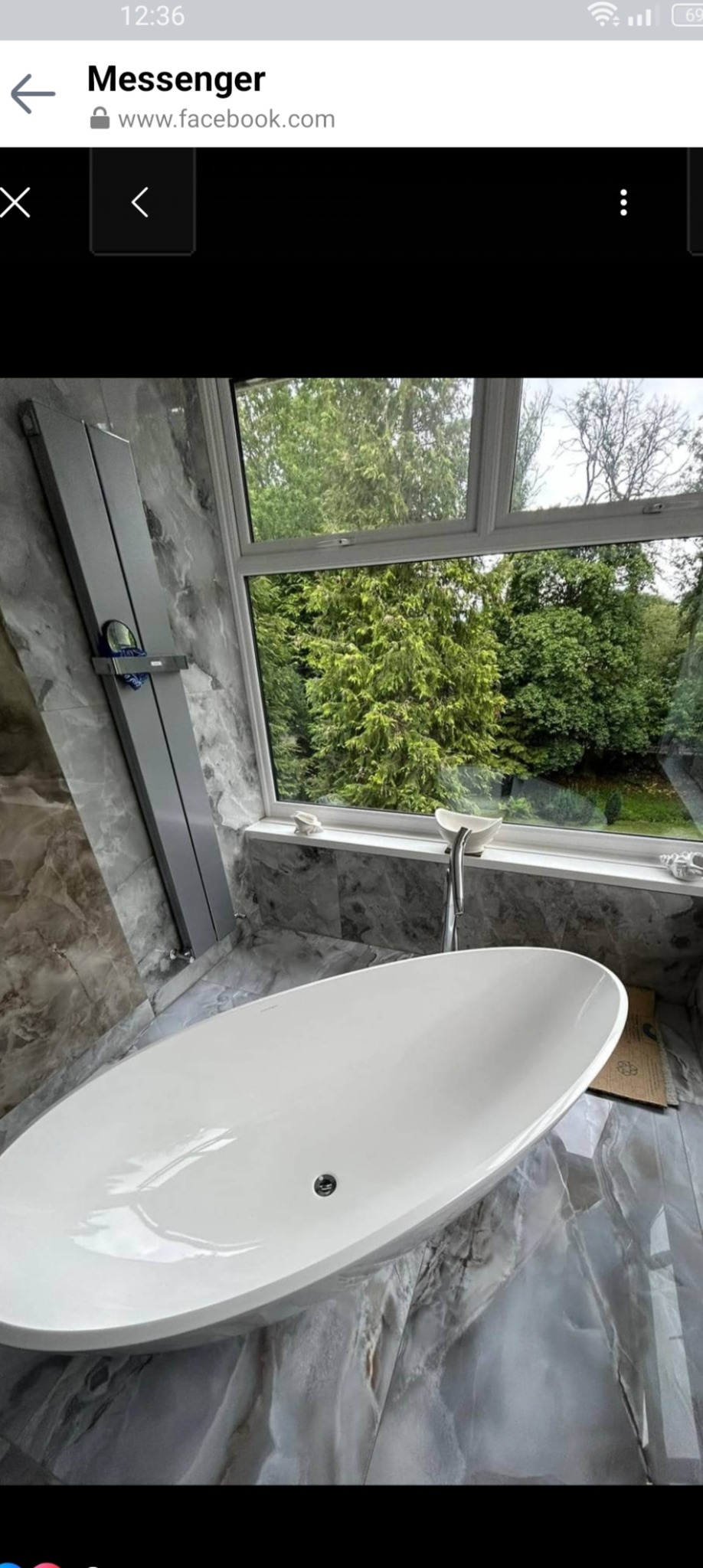 Nowoczesna, wolnostojąca wanna owalna z chromowaną baterią, ustawiona pod oknem z widokiem na zieleń, ściany i podłoga wyłożone płytkami imitującymi marmur w odcieniach szarości, obok wanny leżą...