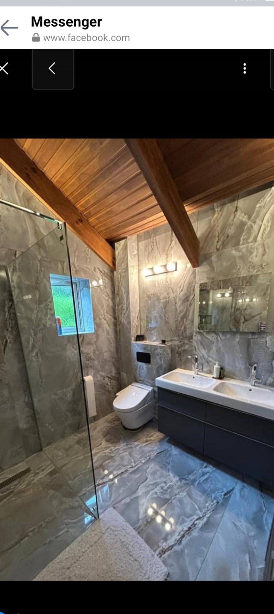 Nowoczesna łazienka z marmurowymi płytkami na ścianach i podłodze, drewnianym sufitem i podwójną umywalką z ciemnoszarą szafką, z widoczną kabiną prysznicową ze szklanymi drzwiami oraz toaletą.