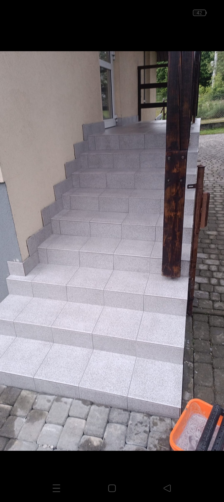 Zewnętrzne schody wyłożone jasnoszarymi płytkami, z drewnianą barierką po prawej stronie, widoczny pojemnik z wodą i narzędzia na dole.