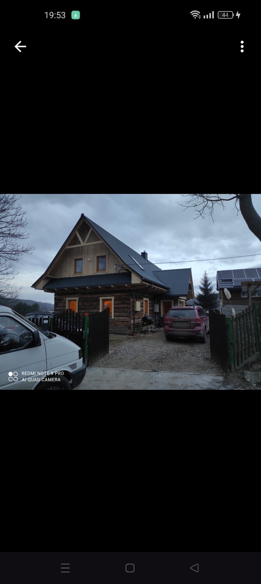 Drewniany dom z ciemnym dachem i oświetlonymi oknami, widoczny zza ogrodzenia, z dwoma zaparkowanymi samochodami i panelem słonecznym na sąsiednim budynku.