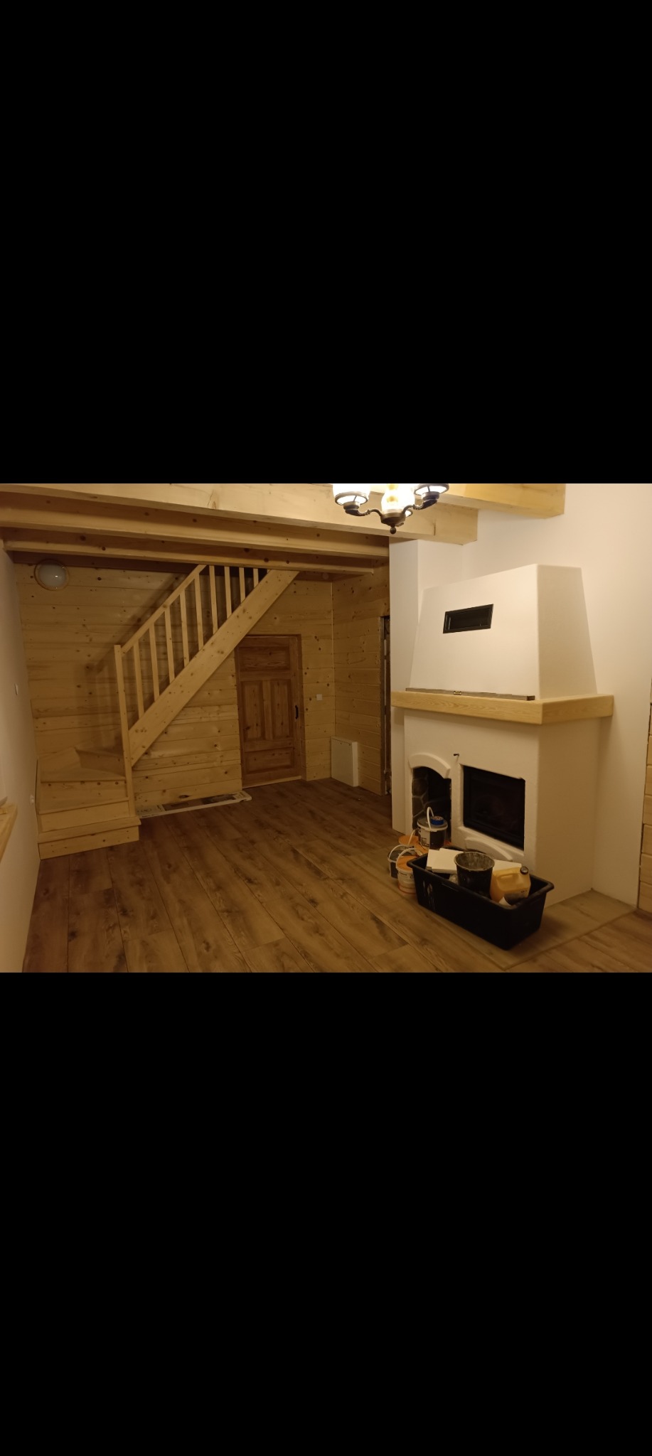 Wnętrze domu z drewnianymi ścianami, schodami i podłogą, biały kominek z czarnym wkładem, widoczne narzędzia malarskie w pojemniku na podłodze.