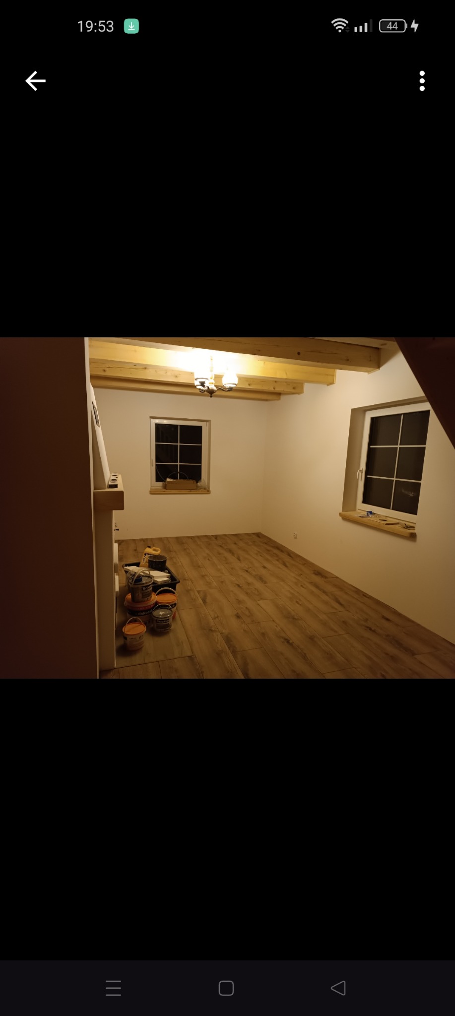 Pomalowane na biało wnętrze poddasza z drewnianymi belkami stropowymi i panelami podłogowymi, widoczne puszki z farbą i narzędzia malarskie.