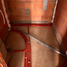 KL Instalacje Konrad Laskowski - Czerwone rury grzewcze w izolacji, szara rura kanalizacyjna, ściany z czerwonej cegły dziurawki, pianka montażowa uszczelniająca, przygotowanie do wylewki pod ogrzewanie podłogowe.