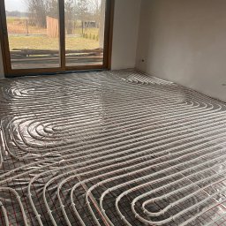 KL Instalacje Konrad Laskowski - Białe rurki ogrzewania podłogowego ułożone spiralnie na folii z czerwoną siatką w surowym pomieszczeniu z dużym oknem na ogród.