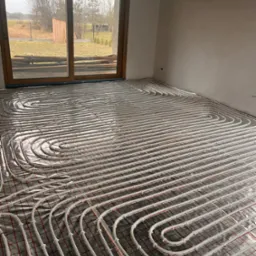 Białe rurki ogrzewania podłogowego ułożone spiralnie na folii z czerwoną siatką w surowym pomieszczeniu z dużym oknem na ogród.