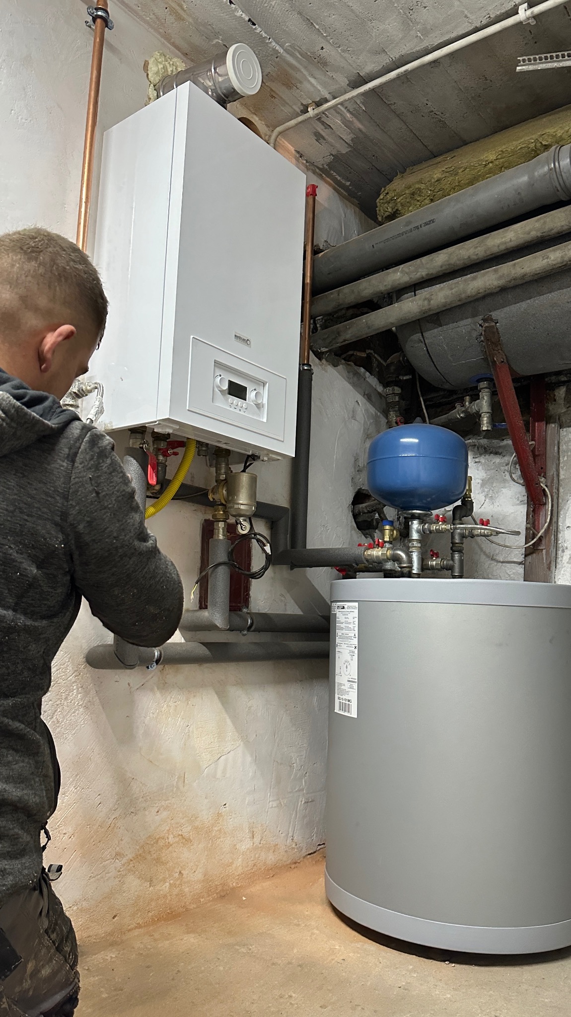 Instalator w szarej bluzie podłącza nowy, biały piec gazowy do instalacji centralnego ogrzewania w piwnicy; widoczny zbiornik wyrównawczy i rury miedziane.