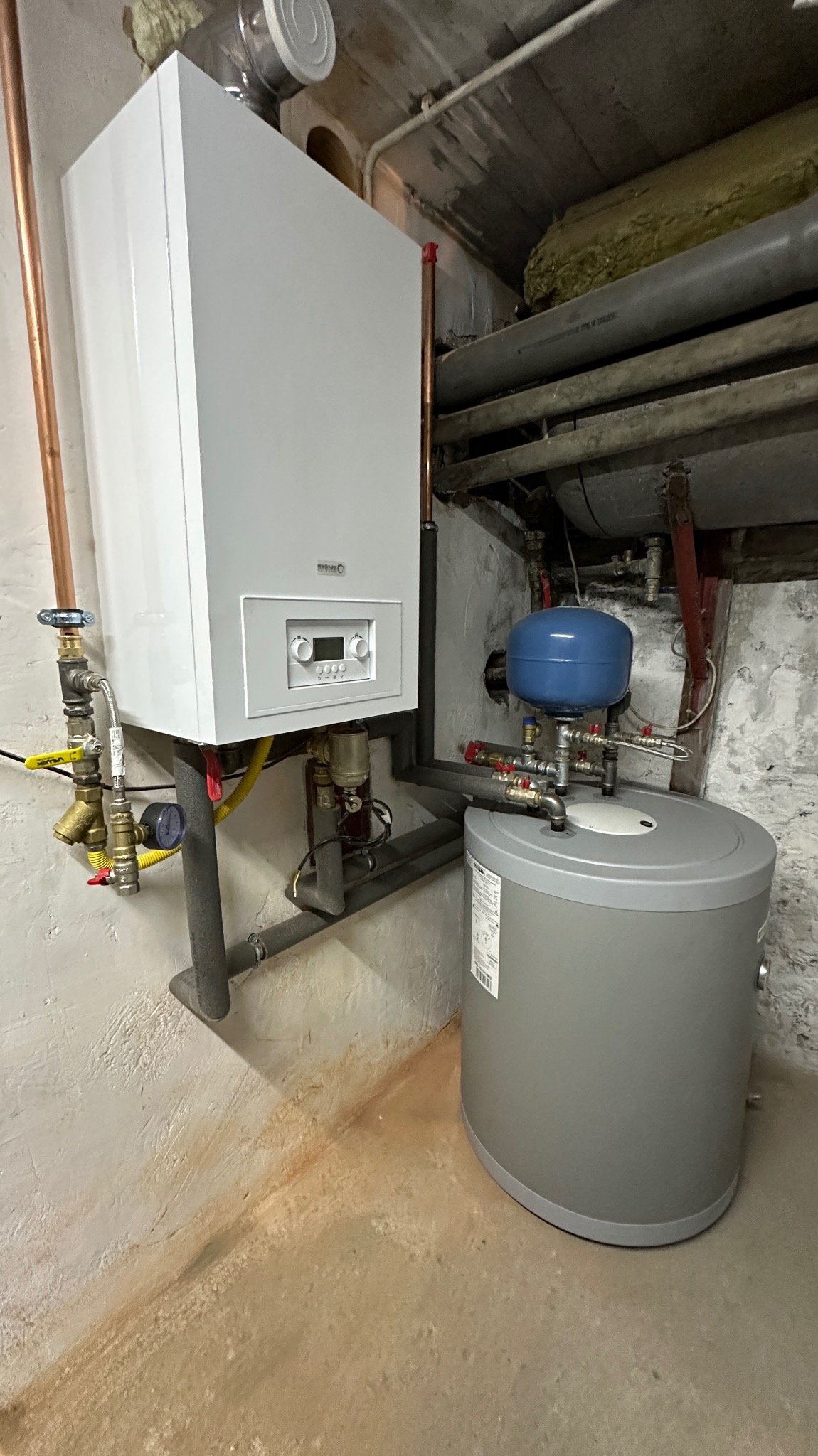 Nowoczesny, biały piec gazowy wiszący na ścianie w piwnicy, z szarym zbiornikiem wyrównawczym i rurami miedzianymi, na tle surowej ściany z widocznymi śladami wilgoci.