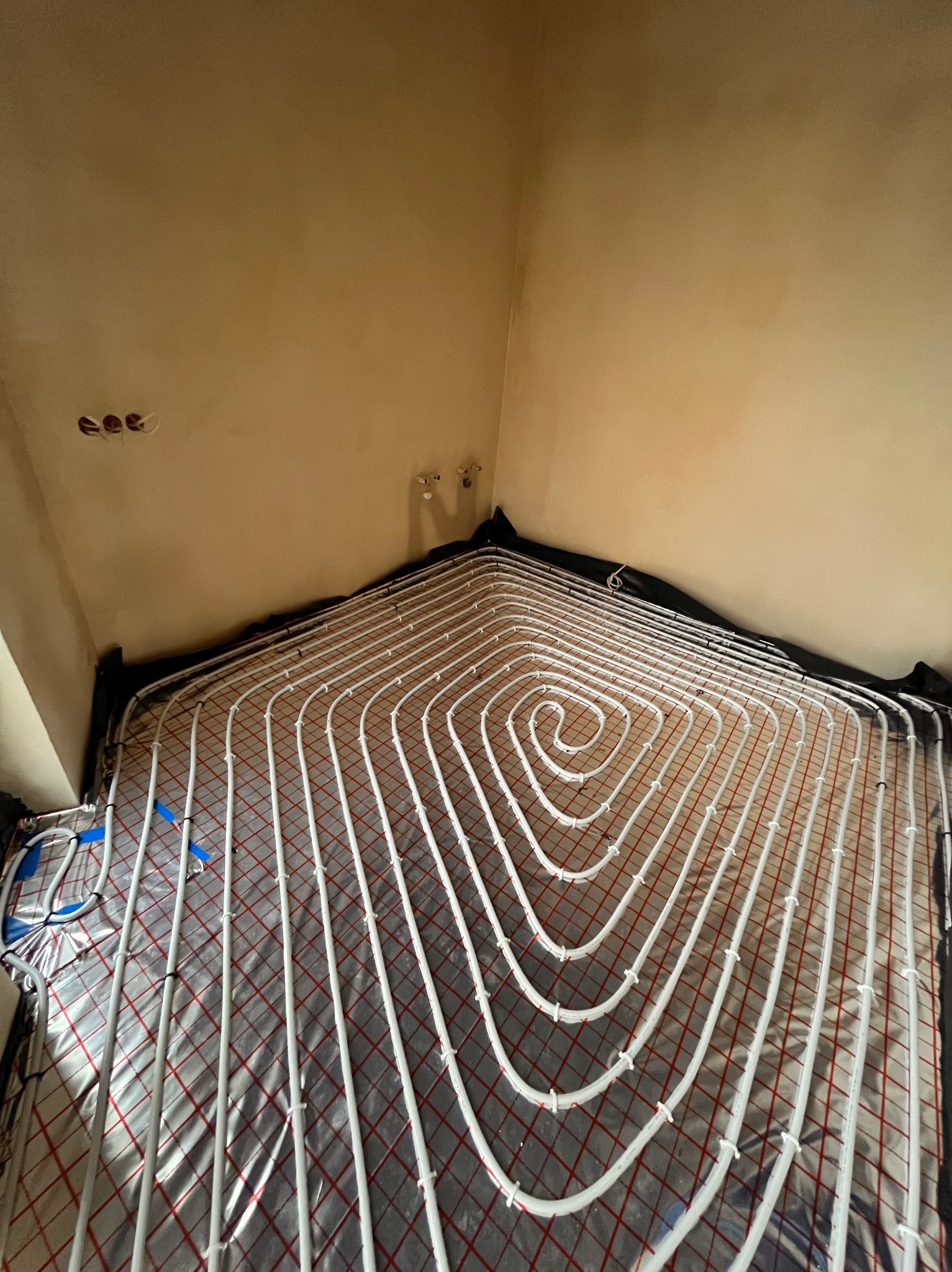 Widok z góry na rozłożone białe rurki ogrzewania podłogowego na folii izolacyjnej z czerwoną siatką, widoczne niepodłączone przewody i puszki elektryczne w tle.