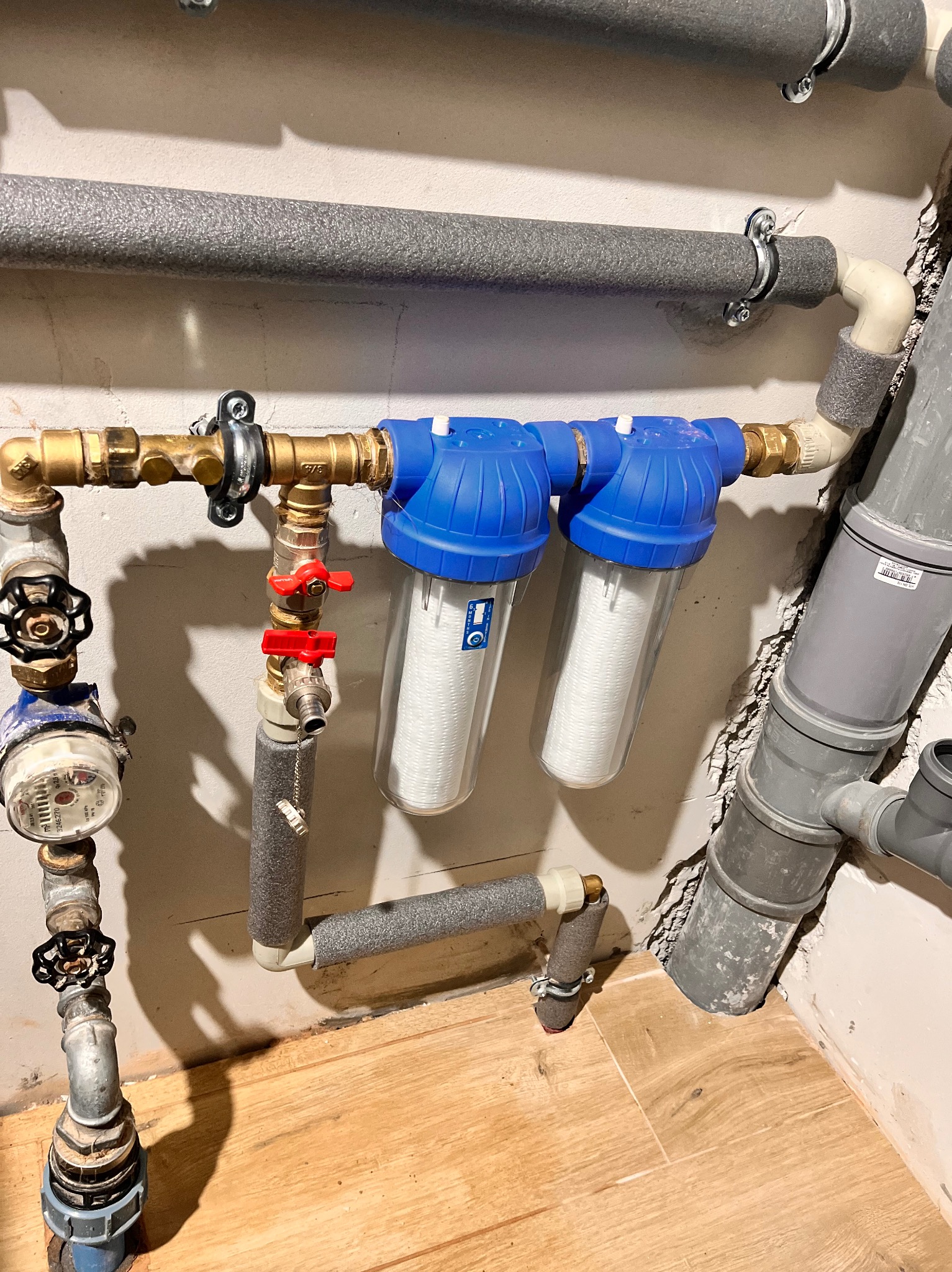 Dwa filtry wody z niebieskimi obudowami zamontowane na miedzianej instalacji, widoczny licznik wody z czarnym zaworem, rury kanalizacyjne z szarego tworzywa sztucznego, całość na tle...