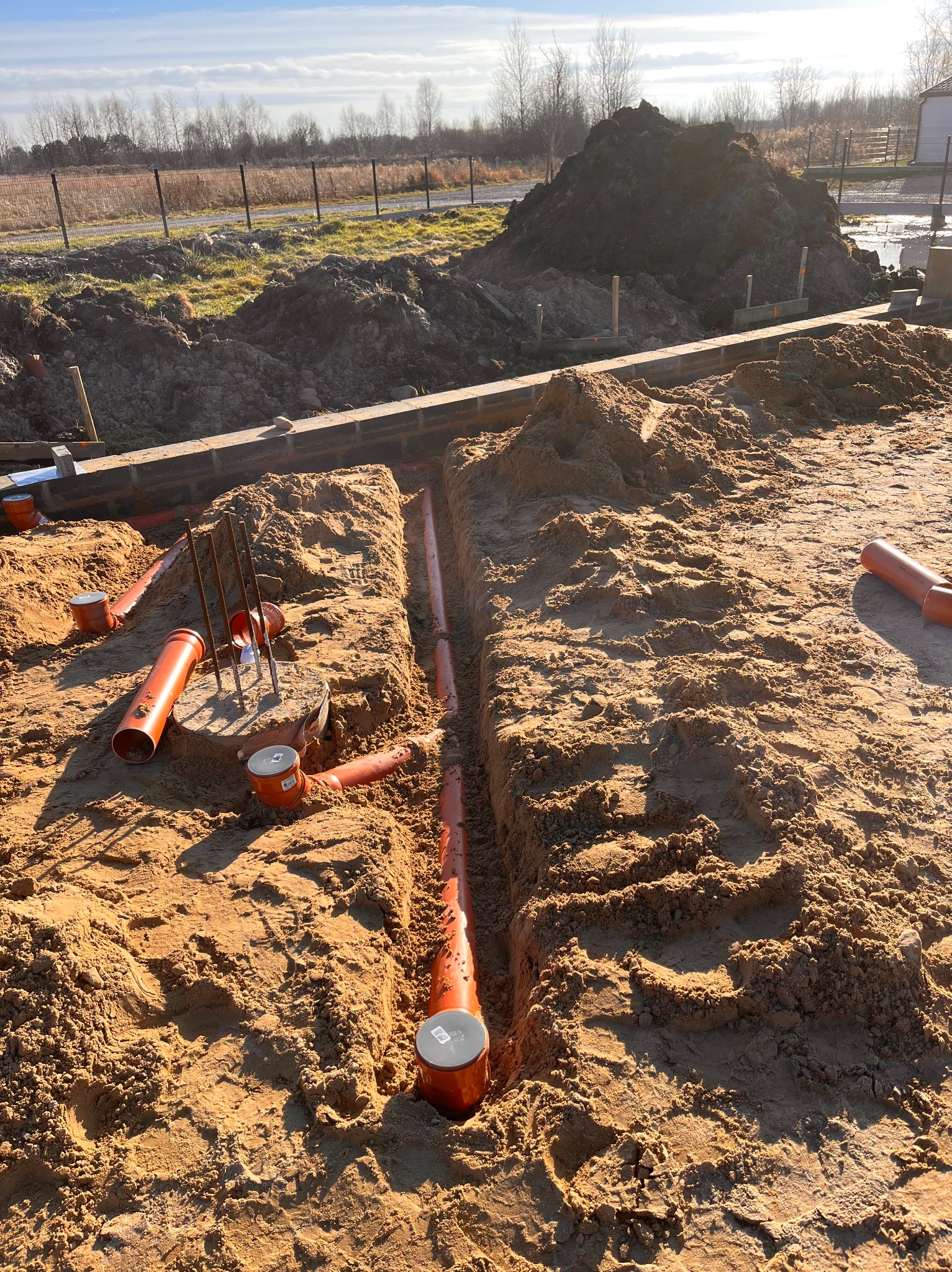 Pomarańczowe rury kanalizacyjne ułożone w wykopie w piasku, widoczne elementy fundamentów i pręty zbrojeniowe, w tle hałda ziemi i krajobraz wiejski.