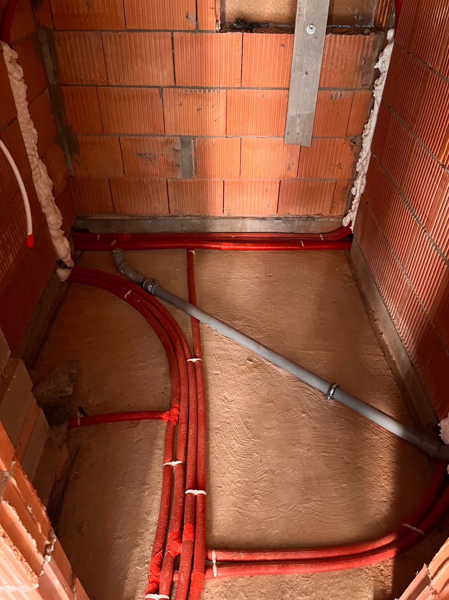 Czerwone rury grzewcze w izolacji, szara rura kanalizacyjna, ściany z czerwonej cegły dziurawki, pianka montażowa uszczelniająca, przygotowanie do wylewki pod ogrzewanie podłogowe.