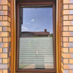 Okno z brązową ramą, moskitierą i plisami, zamontowane w ścianie z cegły. W szybie odbija się budynek z czerwoną dachówką i niebo.