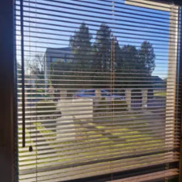 Brązowe żaluzje poziome w oknie z widokiem na ogród, budynek i błękitne niebo w słoneczny dzień. Regulacja światła za pomocą żaluzji.