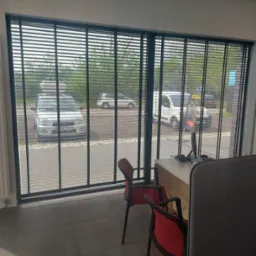 Ciemne żaluzje poziome w oknie biura, widok na ulicę z samochodami. Dwa krzesła biurowe w kolorze czerwonym w pomieszczeniu.