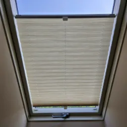 Beżowa plisa okienna zamontowana w oknie dachowym, widok od dołu. Częściowo zasłonięte okno z widocznym fragmentem nieba. Uchwyt okienny w dolnej części ramy.