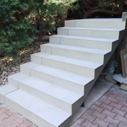 Wykończone płytkami schody zewnętrzne z jasnego gresu, widok z dołu, na pierwszym planie wiadro i kielnia, w tle zielona roślinność.