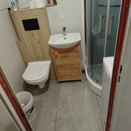 ANDRII KUN - Ukończona łazienka z kabiną prysznicową, umywalką z szafką z drewnopodobnym fornirem, podwieszaną toaletą i pralką, widoczna przez futrynę drzwi.