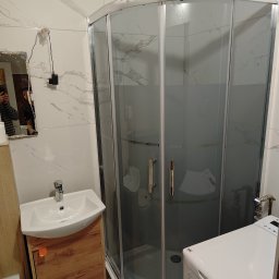 ANDRII KUN - Umywalka z szafką z drewnopodobnym frontem, kabina prysznicowa narożna z przesuwnymi drzwiami oraz pralka w małej łazience wykończonej białymi płytkami imitującymi marmur.