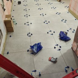 ANDRII KUN - Układanie jasnych płytek podłogowych w trakcie remontu, z użyciem poziomicy, systemu poziomowania płytek i paczek z akcesoriami widocznych na podłodze.