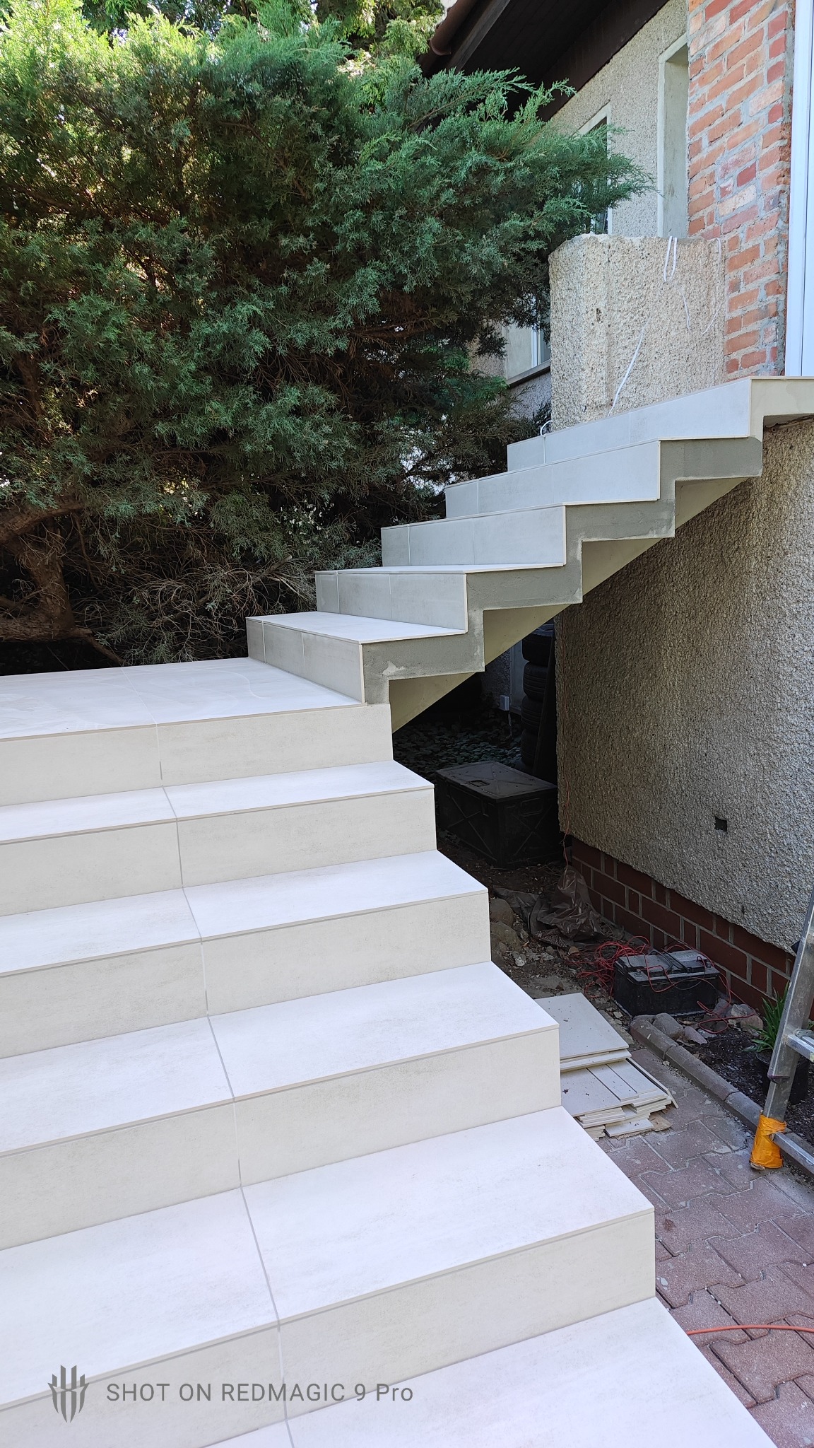 Zewnętrzne schody wyłożone jasnymi płytkami ceramicznymi, widoczne ślady fugowania, w tle bujna zieleń i fragment elewacji budynku.