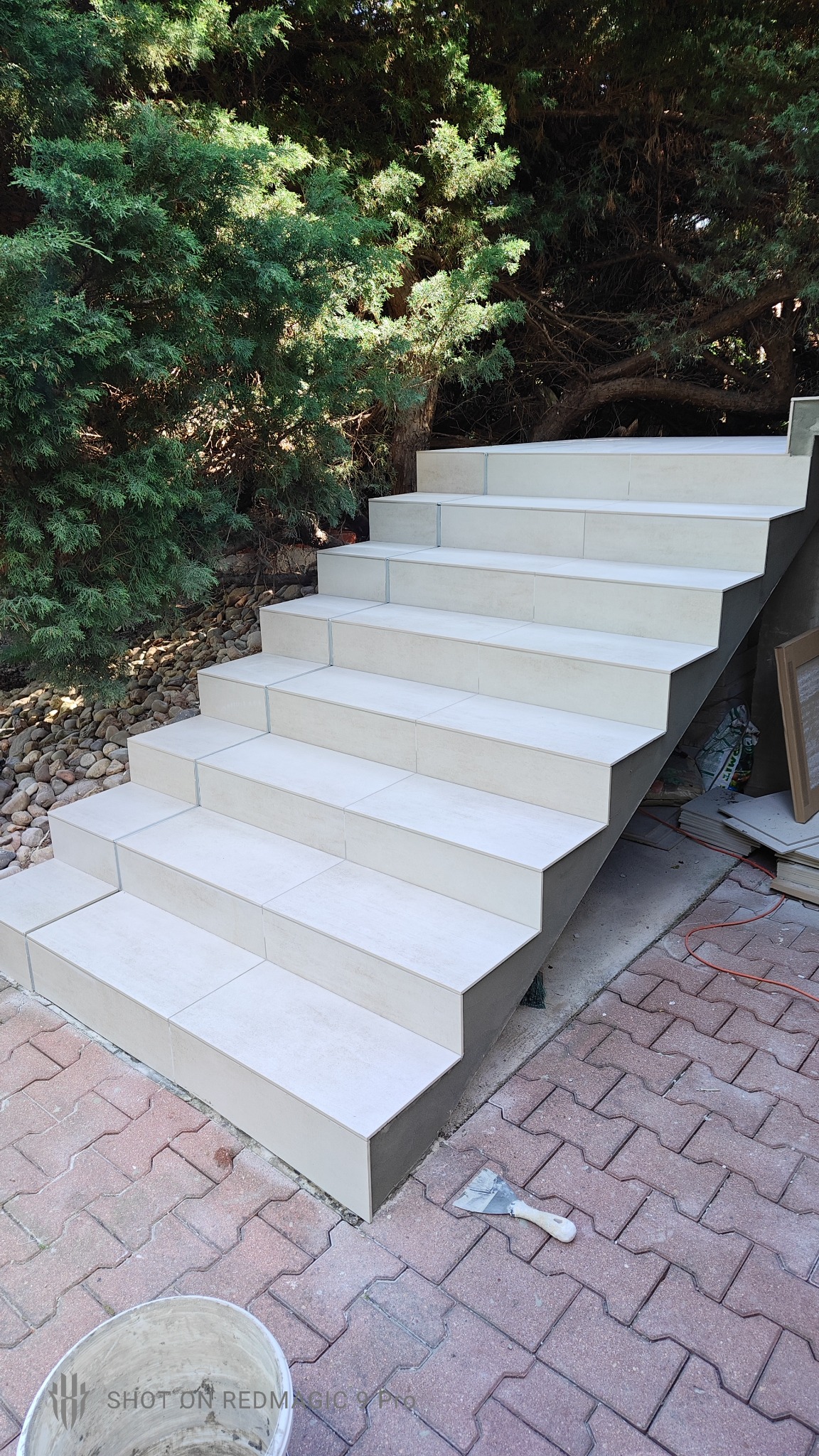 Wykończone płytkami schody zewnętrzne z jasnego gresu, widok z dołu, na pierwszym planie wiadro i kielnia, w tle zielona roślinność.