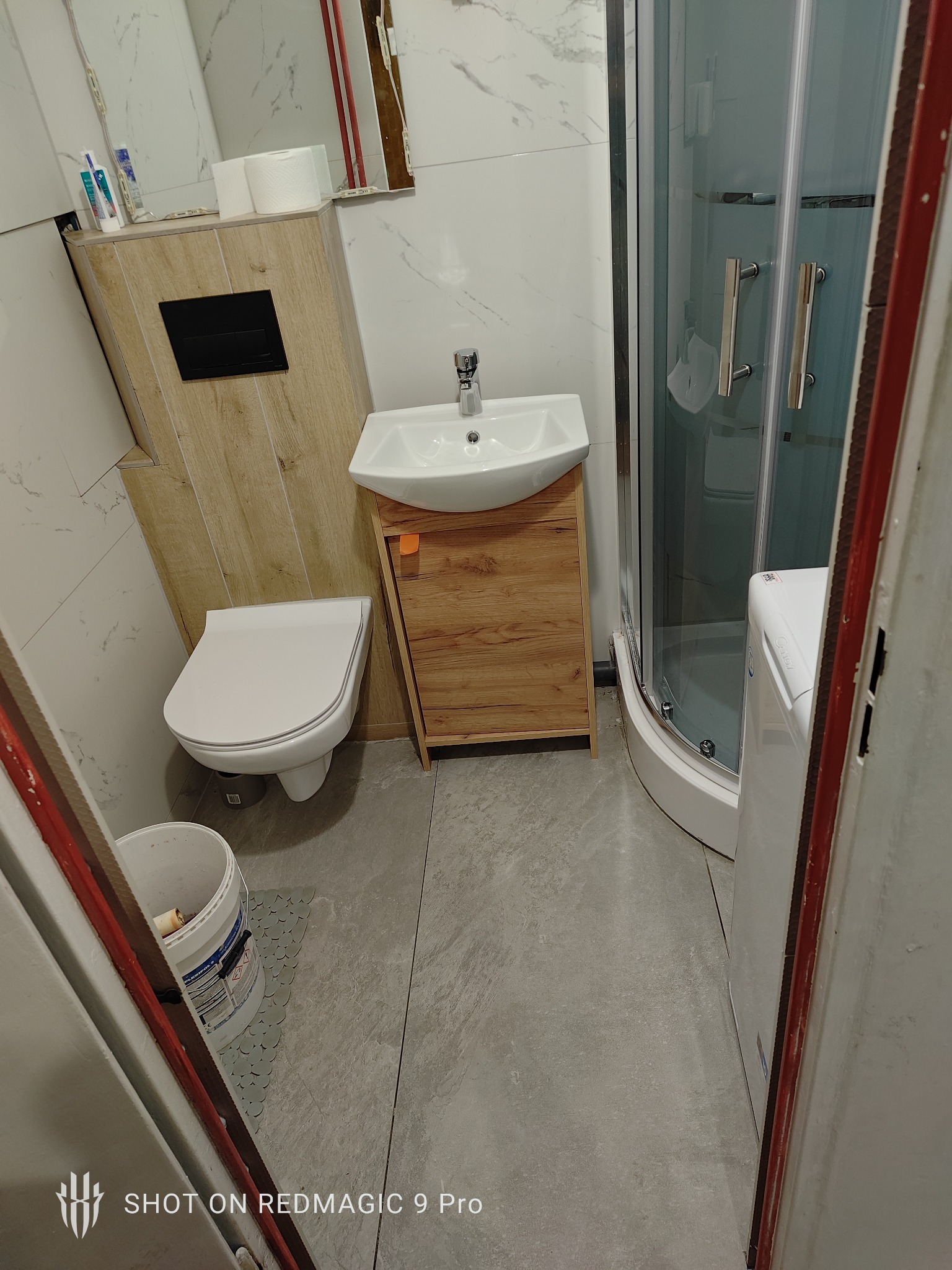 Ukończona łazienka z kabiną prysznicową, umywalką z szafką z drewnopodobnym fornirem, podwieszaną toaletą i pralką, widoczna przez futrynę drzwi.