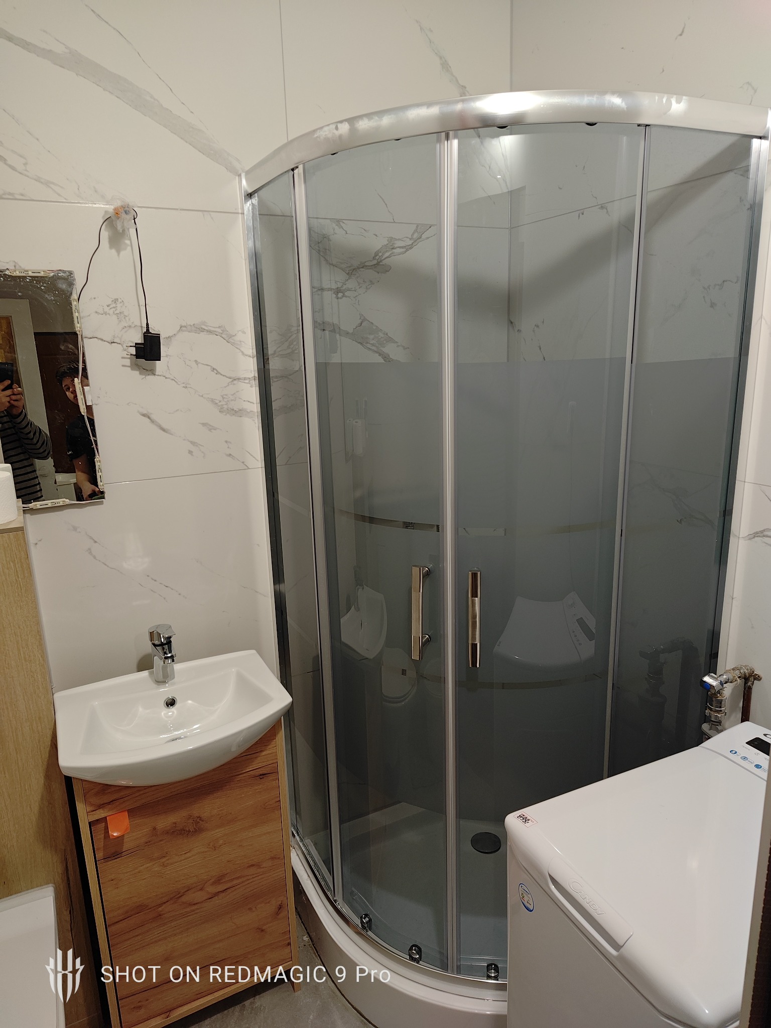 Umywalka z szafką z drewnopodobnym frontem, kabina prysznicowa narożna z przesuwnymi drzwiami oraz pralka w małej łazience wykończonej białymi płytkami imitującymi marmur.