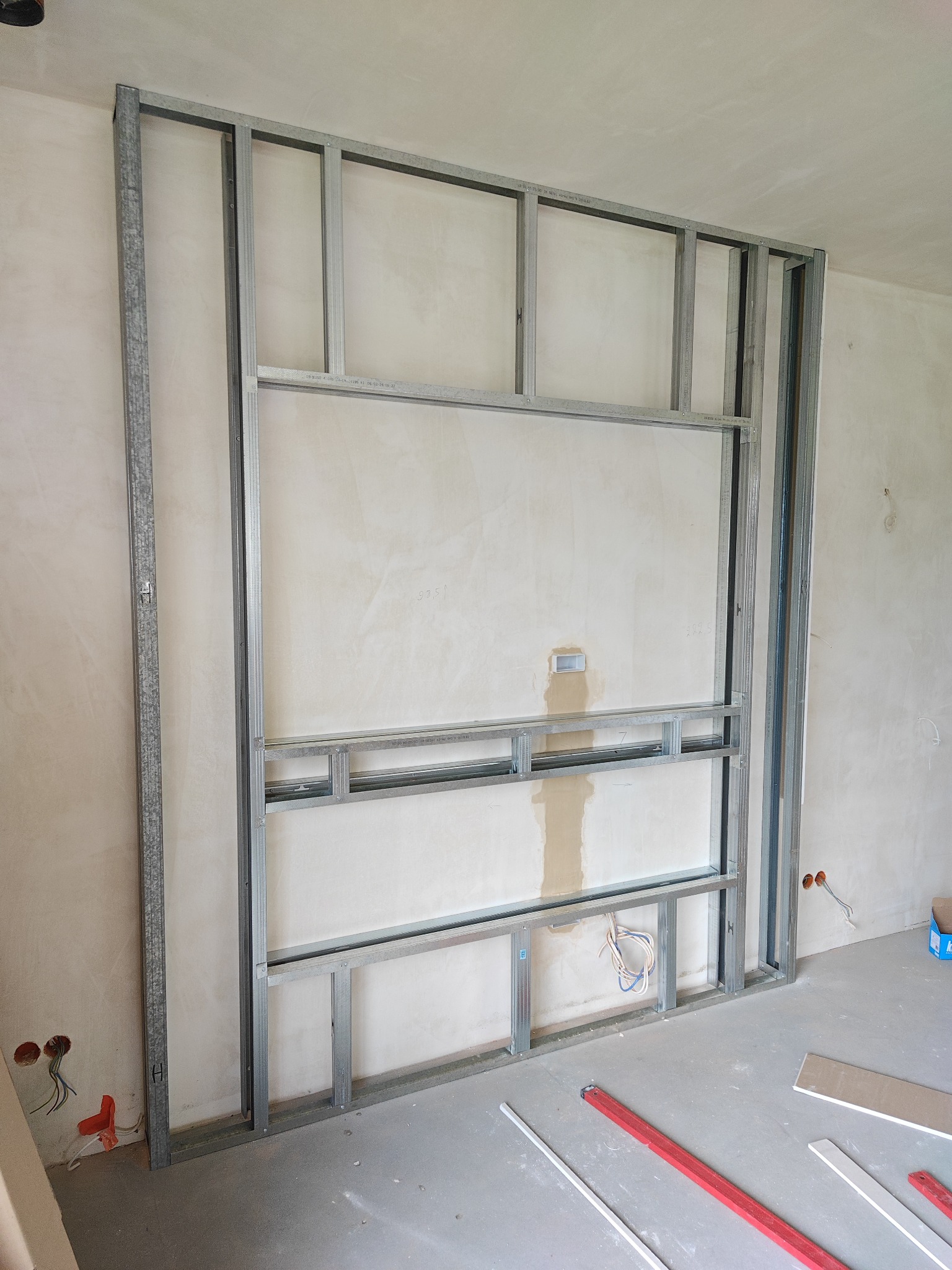 Stelaż z profili metalowych do zabudowy ściany działowej, widoczne przewody elektryczne, elementy poziomicy i płyty gipsowej na podłodze.