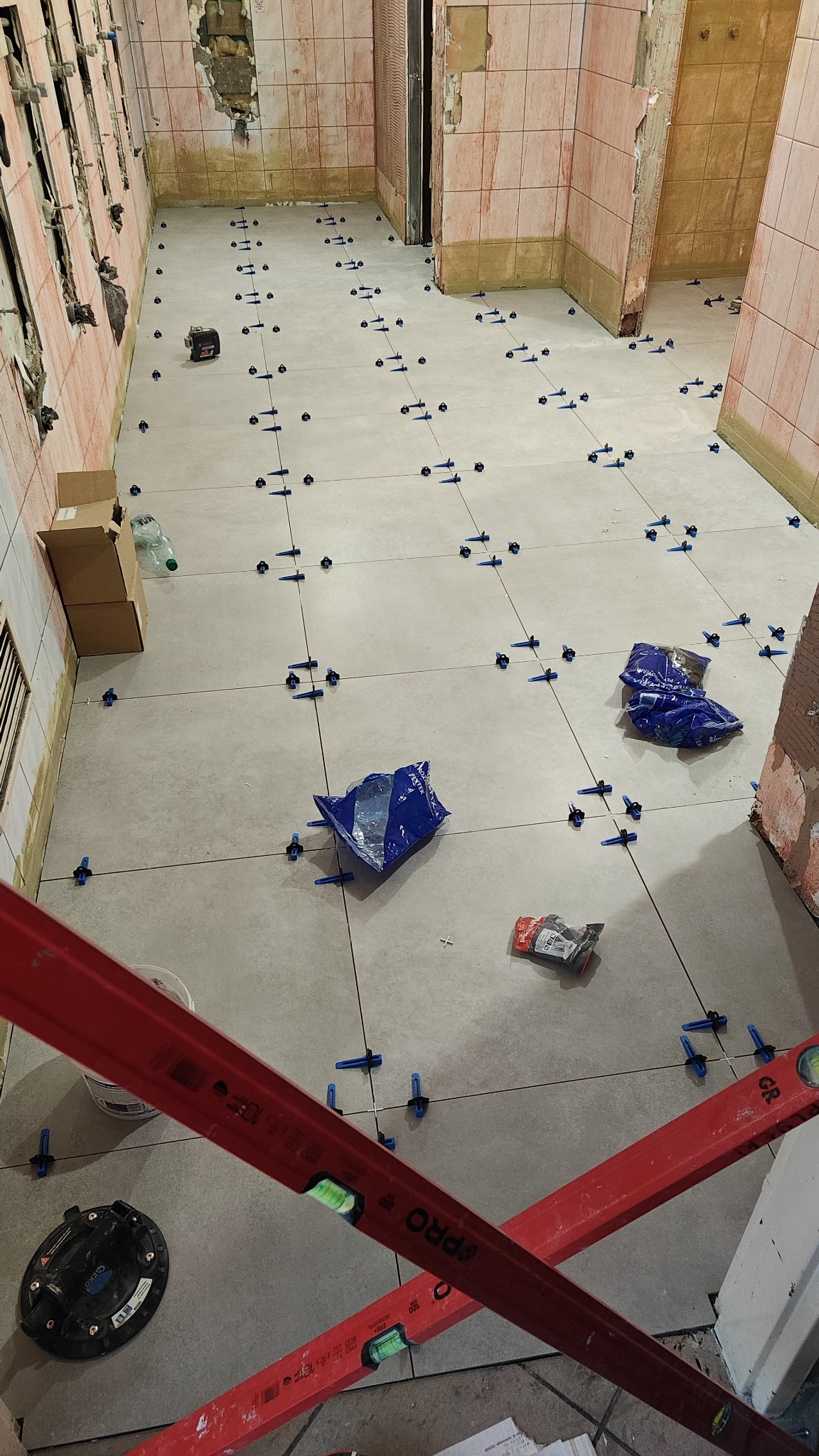 Układanie jasnych płytek podłogowych w trakcie remontu, z użyciem poziomicy, systemu poziomowania płytek i paczek z akcesoriami widocznych na podłodze.