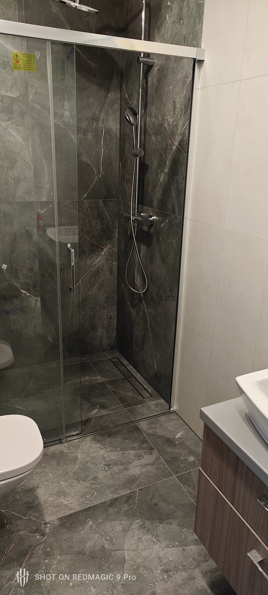 Nowoczesna kabina prysznicowa z ciemnymi płytkami imitującymi marmur i odpływem liniowym, widoczna toaleta i umywalka z szafką w łazience po remoncie.