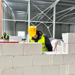 AT-BUD - Murarz w żółtym kasku i kamizelce, w trakcie murowania ściany z białych bloczków betonu komórkowego wewnątrz hali, z widocznym sznurkiem murarskim i poziomnicą, drugi pracownik w tle.