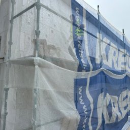 AT-BUD - Budynek w trakcie ocieplania elewacji, widoczne rusztowania osłonięte siatką z reklamą, częściowo zasłaniającą ściany z bloczków betonu komórkowego.