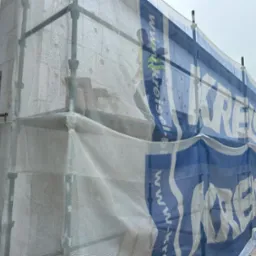 Budynek w trakcie ocieplania elewacji, widoczne rusztowania osłonięte siatką z reklamą, częściowo zasłaniającą ściany z bloczków betonu komórkowego.