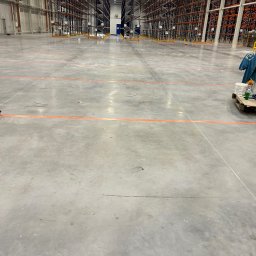 AT-BUD - Wnętrze pustego magazynu z betonową podłogą z pomarańczowymi liniami, metalowymi regałami w tle i paletą z farbami oraz ubraniem roboczym po prawej stronie.