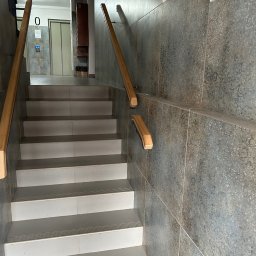 AT-BUD - Klatka schodowa w budynku z wykończonymi schodami z płytek i drewnianą balustradą, widok na korytarz z windą.