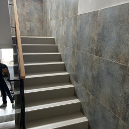 AT-BUD - Wykończone schody wewnętrzne z szarobrązowymi płytkami dekoracyjnymi na ścianie i jasnymi stopniami, widoczny fragment poręczy i osoba w tle.