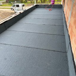 AT-BUD - Wykończony płaski dach pokryty papą termozgrzewalną, widoczny gazowy palnik z butlą propan-butan, ściana budynku w tle.