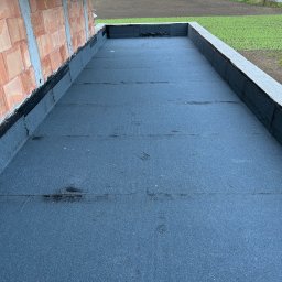 AT-BUD - Wykończony płaski dach z papy termozgrzewalnej, widok z góry, surowa ściana budynku w tle, za nią pola i drzewa.