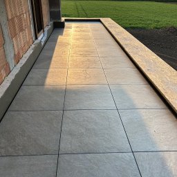 AT-BUD - Świeżo ułożone szare płytki na tarasie domu w trakcie budowy, widoczne cegły i ocieplenie, w tle pole i zachodzące słońce.