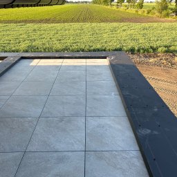 AT-BUD - Szare płytki tarasowe ułożone na zewnątrz budynku, z widokiem na pole uprawne w tle, czarne obramowanie z widocznymi śladami butów.